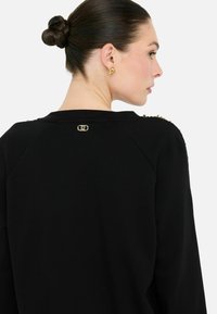 Zwarte sweatshirt met een gladde textuur, voorzien van gouden hardware accenten op de schouders en een logo op de achterkant van de halslijn.