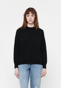 Pull noir à col haut, à coupe ample et épaules tombantes, associé à un jean clair déchiré.