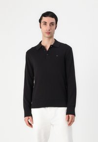 Černé polo tričko s dlouhým rukávem a límečkem, s knoflíkovou légou na tři knoflíky, žebrovaným lemem a manžetami, se malou logovou aplikací na hrudi.