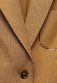 Gros plan sur les revers d'un blazer marron et un seul bouton marron foncé avec une texture de tissu visible et des coutures.