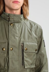 Olivově zelená bunda s vysokým límcem, která má několik kapes, bronzové knoflíky a zipovou kapsu na boku. Texturovaný povrch látky.