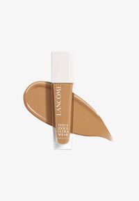Lancôme Teint Idole Ultra Wear foundation i en klar glassbeholder med hvit lokk. Rikt, middels brunt væske med en jevn tekstur samlet ved siden av.