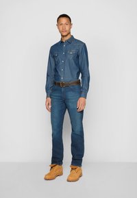 Chemise en denim bleu avec un empiècement pointu, deux poches poitrine, associée à un jean bleu foncé et des bottes à lacets marron clair. Une ceinture marron ajoute du contraste.