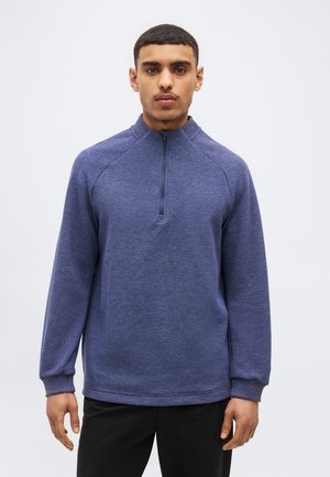 Moški s kratkimi lasmi, oblečen v modri pulover z dolgimi rokavi in četrtinskim zadrgo ter črne hlače, na svetlem enobarvnem ozadju.