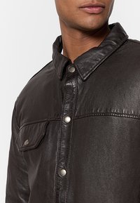 Schwarze Lederjacke mit spitzem Kragen, Druckknöpfen und einer Brusttasche; glatte Textur mit leicht glänzender Oberfläche.