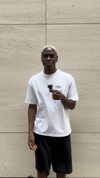 Camiseta blanca con un pequeño gráfico en el pecho izquierdo, pantalones cortos negros, sosteniendo un micrófono con una pantalla antiviento esponjosa frente a una pared texturizada.