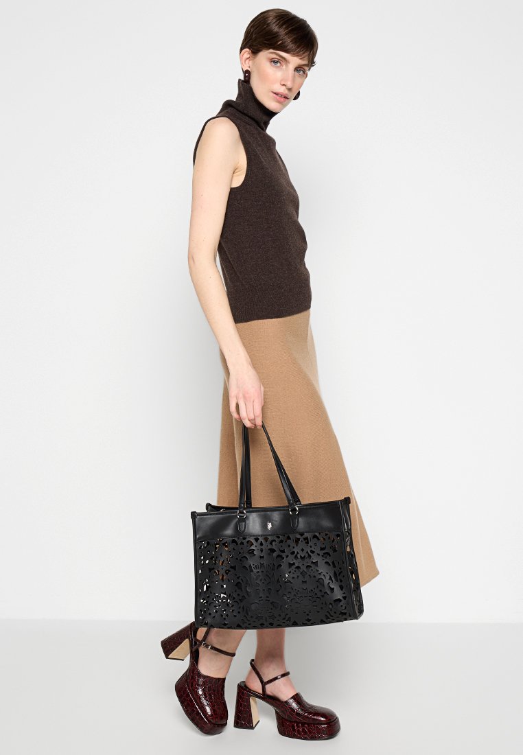 Bolso tote de cuero negro con patrón floral cortado a láser, asas planas y forma estructurada. El modelo lleva una blusa sin mangas marrón y una falda beige.