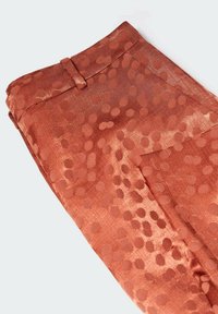 Pantalon couleur rouille avec un motif à pois subtil, plié soigneusement avec la ceinture et les passants visibles sur un fond uni.