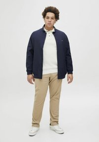 Chaqueta ligera azul marino con cremallera en la parte delantera, llevada sobre un suéter de punto blanco. Pantalones beige y zapatillas blancas completan el look. Diseño simple, sin patrones.