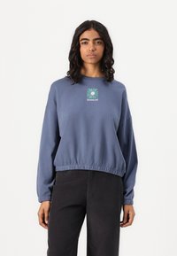 Mėlynas trumpas džemperis su elastinėmis rankovėmis ir apačia. Papuoštas žaliu ir baltu grafiniu spaudiniu, pažymėtu "QUIKSILVER". Malonus ir lygus paviršius.