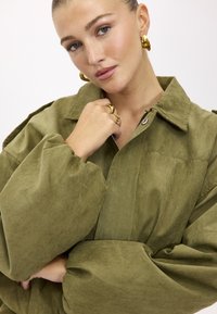 Jeune femme aux yeux bleus portant une veste oversize vert olive et des boucles d'oreilles créoles en or, avec des bagues, posant la main près du visage.