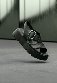 Black platform sandals with cross straps made of smooth leather, featuring a metallic buckle and a chunky, textured sole.