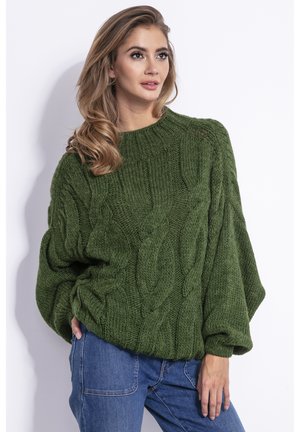Zielony sweter w splot kablowy z wysokim dekoltem, oversize'owy fason i gruba tekstura. Połączony z niebieskimi dżinsami. Minimalistyczne tło.