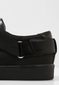 Baskets noires avec un bout en cuir lisse, une tige en nylon texturé, une sangle élastique ajustable et un détail logo sur le côté.