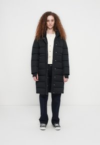 Μαύρο μπουφάν puffer με χαλαρή εφαρμογή, διακοσμημένο με ψηλό γιακά, κουμπιά κλιπ και οριζόντιο καπιτονέ σχέδιο. Συνδυασμένο με σκούρο τζιν και αθλητικά παπούτσια.