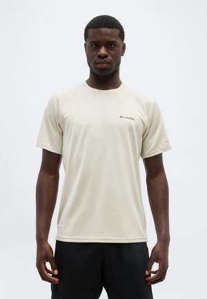 Ενήλικας άνδρας που στέκεται, φοράει ανοιχτό μπεζ κοντομάνικη μπλούζα Columbia και μαύρο παντελόνι, με ουδέτερη έκφραση προσώπου.