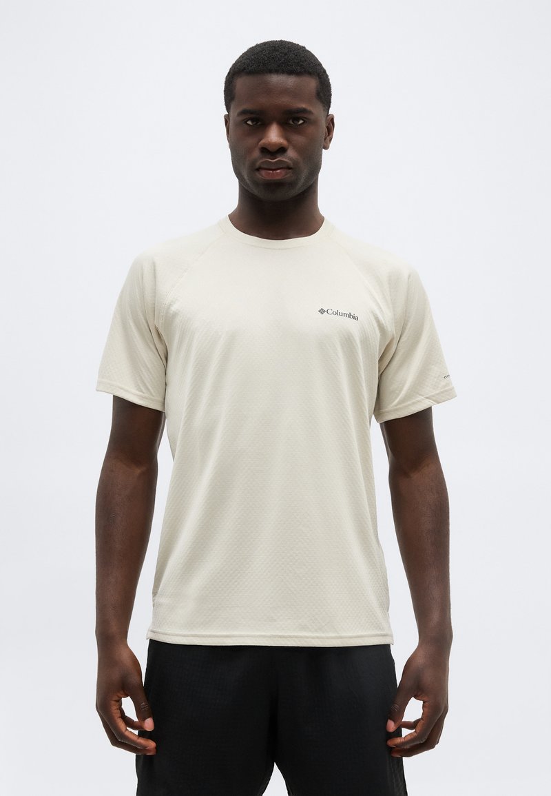 Ενήλικας άνδρας που στέκεται, φοράει ανοιχτό μπεζ κοντομάνικη μπλούζα Columbia και μαύρο παντελόνι, με ουδέτερη έκφραση προσώπου.