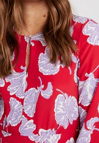 Vestido rojo con grandes patrones florales en lila, escote redondo con detalle de ojal y mangas largas con una textura suave.
