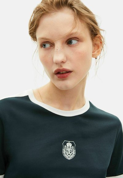 Dunkelgrünes Baumwoll-T-Shirt mit weißem Kragen, ausgestattet mit einem grafischen Emblem auf der Brust, glatte Textur und lässigem Design.
