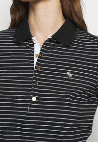Černobílý pruhovaný polo tričko s černým límečkem, třemi zlatými knoflíky a malým vyšitým logem na hrudi.