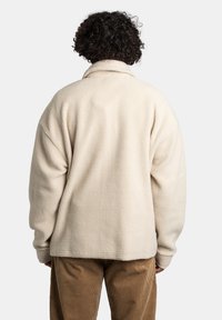 Jasno kremowa polarowa kurtka o miękkiej teksturze, luźnym kroju i kołnierzu. Pokazana z tyłu, z opadającymi ramionami i dolnym brzegiem, który sięga poniżej talii.