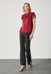 Rød bluse med rynker og nøkkelhulldetalj, matchet med svarte vide bukser. Modellen har på seg svarte sandaler med stropper. Glatte teksturer gjennom hele antrekket.