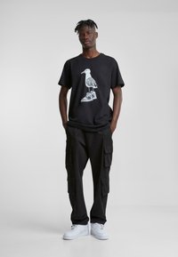 Sort bomuld t-shirt med et hvidt fuglemotiv. Sorte cargo-bukser med flere lommer. Hvide sneakers. Ensfarvet grå baggrund.