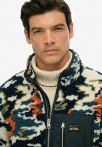 Chaqueta de forro polar con un patrón abstracto multicolor, que presenta un cuello alto, cierre con cremallera en la parte delantera y un bolsillo en el pecho con un parche de logo negro.
