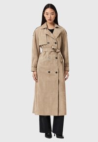 Μπεζ καμπαρντίνα από σουέντ με διπλό πέτο, ζώνη και πλαϊνές τσέπες. Διαθέτει γιακά και μανσέτες με κουμπιά. Συνδυασμένη με μαύρο παντελόνι.