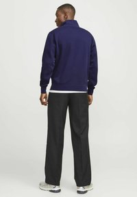 Sudadera de cuello con cremallera en azul marino con puños acanalados, combinada con pantalones de pierna ancha negros. El modelo se encuentra de pie de perfil a la izquierda contra un fondo claro.