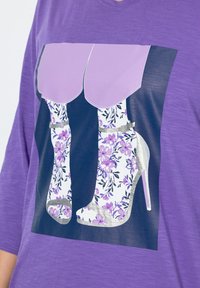 Lila T-shirt med en grafik av blommönstrade högklackade skor med silverdetaljer. Designen kontrasterar med tröjans textur och färg.