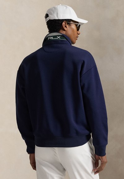 Φούτερ σε ναυτικό μπλε με ψηλό γιακά, διακοσμημένο με πράσινο και λευκό λογότυπο RLX. Μαλακή υφή και ριμώδη μανσέτες στα μανίκια και στο τελείωμα.