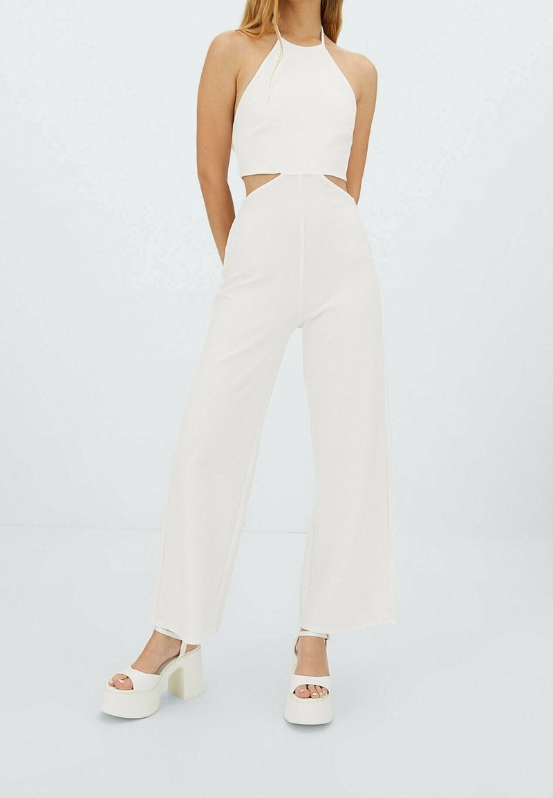 Top corto blanco con cuello halter combinado con pantalones de tiro alto. Tela suave con detalles de cortes y piernas amplias. La modelo lleva tacones altos.