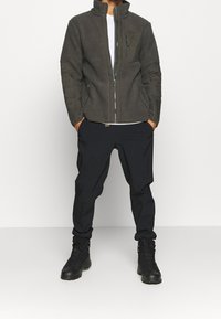 Mężczyzna w ciemnoszarej polarowej kurtce z zamkiem błyskawicznym, czarnych spodniach i czarnych butach turystycznych, stojący na białym tle.