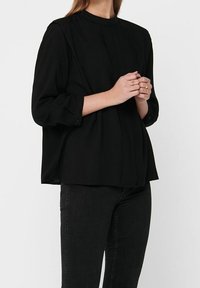 Blouse noire à manches longues avec un col montant, en tissu doux, design plissé et poignets boutonnés, associée à un jean foncé.