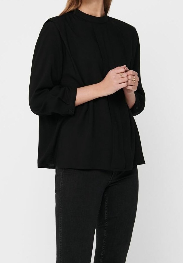 Blouse noire à manches longues avec un col montant, en tissu doux, design plissé et poignets boutonnés, associée à un jean foncé.