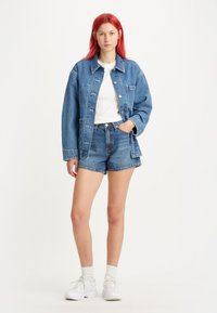 Giacca di jeans blu con grandi bottoni, t-shirt bianca, pantaloncini di jeans a vita alta e sneakers bianche. Texture liscia, vestibilità casual.