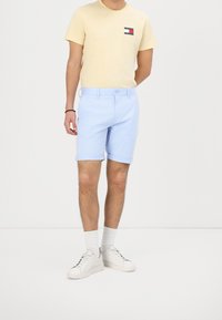 Camiseta amarilla clara con un pequeño logo, combinada con pantalones cortos azul claro. El modelo lleva zapatillas blancas y calcetines cortos, de pie sobre un fondo neutro.