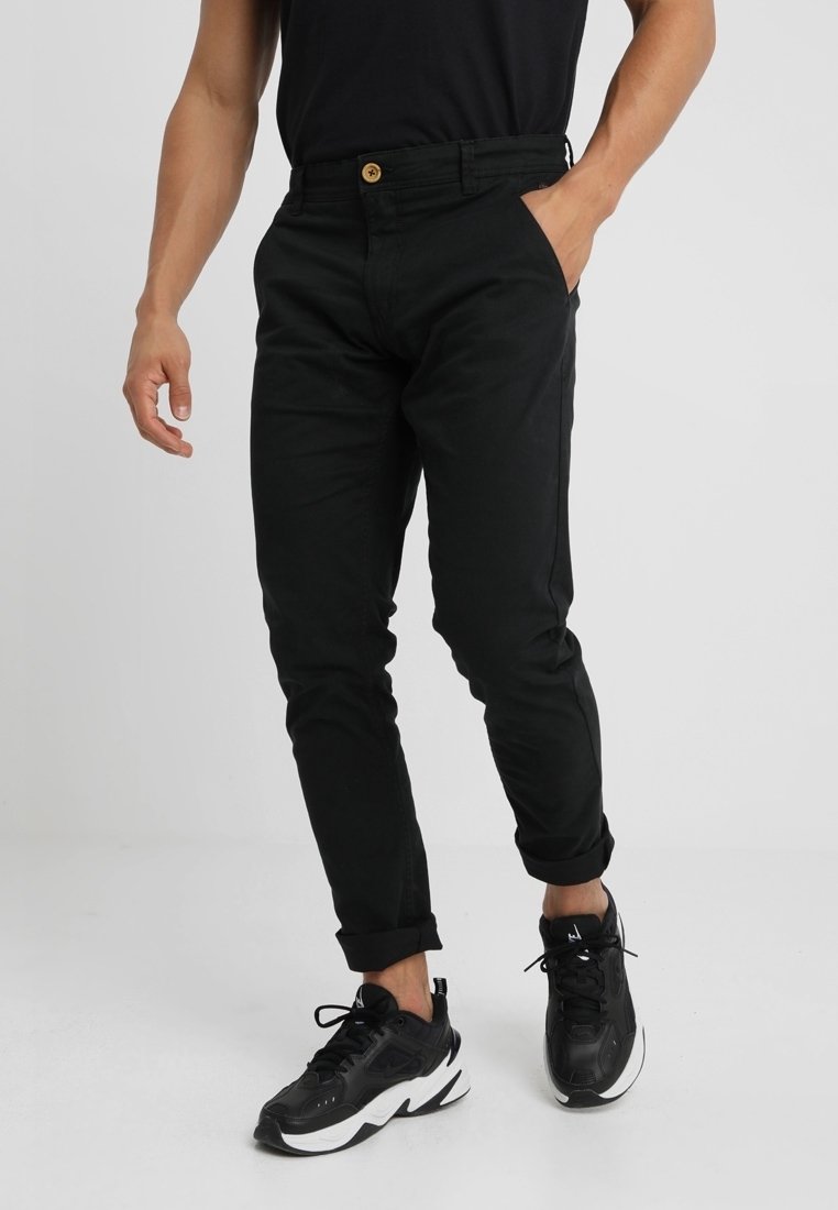 Homme portant un pantalon noir slim avec fermeture à boutons et des baskets noires et blanches, debout avec une main dans la poche.