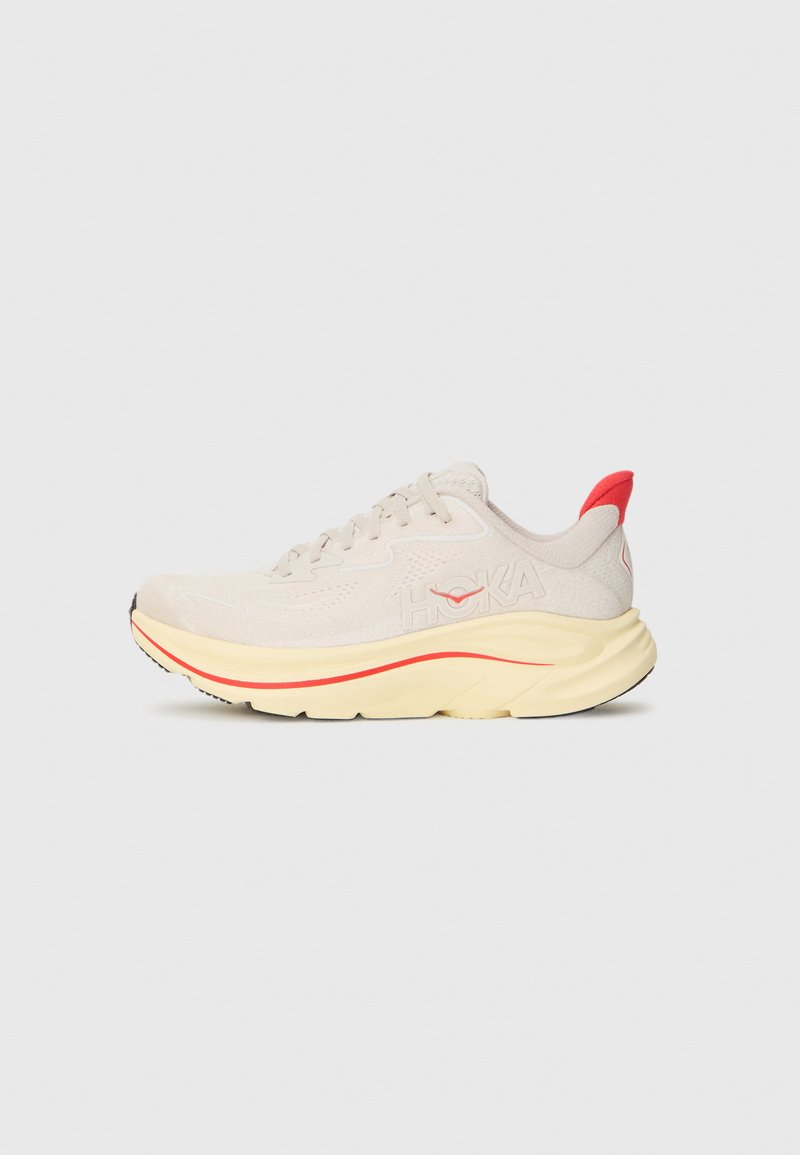Chaussure de sport beige avec une tige en maille texturée, semelle intermédiaire jaune rembourrée et accent rouge au talon. Logo affiché sur le côté.