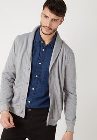 Cárdigan gris con cuello de chal, cierre de botones, tejido suave, usado sobre una camisa de botón azul oscuro con un bolsillo en el pecho. Pantalones negros visibles.