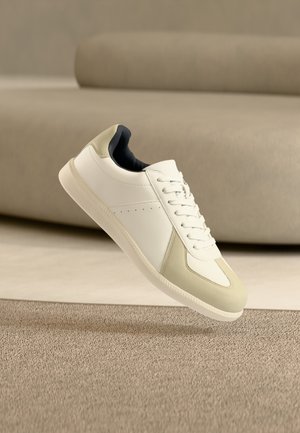 Baskets blanches et beiges à lacets flottant au-dessus d'un tapis beige texturé dans un cadre de pièce minimaliste.