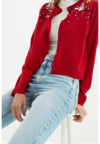 Cardigan rosso con pupazzi di neve e alberi ricamati, dotato di scollo rotondo, chiusura con bottoni e polsini a coste. Abbinato a jeans azzurri.