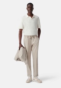 Licht beige poloshirt met korte mouwen, broek met elastische tailleband en trekkoord, lichtgrijze blazer en beige sneakers met off-white accenten.