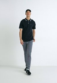 Černé polo tričko s bílými límečkovými akcenty, šedé kalhoty a černé tenisky. Model stojí na neutrálním bílém pozadí.
