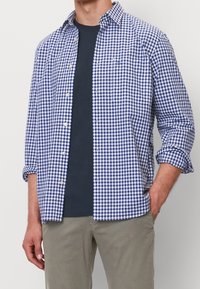 Blauw-wit geruite shirt met button-down kraag, opgerolde mouwen en een borstzak, gelaagd over een donkerblauw T-shirt.