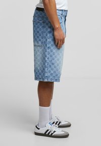 Lysblå denimshorts med et cirkulært mønster, sidelommer og knælangt snit, parret med hvide sokker og sort-hvide sneakers.