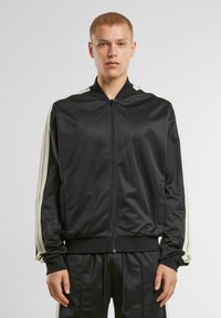 Veste bomber noir en satin avec un col haut, fermeture éclair sur toute la longueur et rayures crème contrastantes le long des manches. Texture lisse, coupe athlétique.