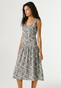 Mujer con vestido sin mangas hasta la rodilla con patrón floral verde y marrón, posando frente a un fondo claro y sencillo.