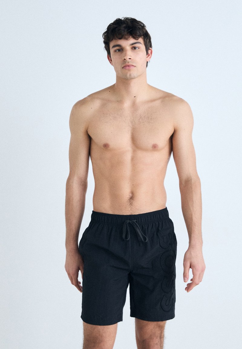 Shorts de bain noirs avec une taille élastique et un cordon de serrage réglable. Comprend des poches latérales et un logo texturé sur la jambe.